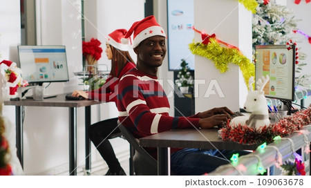 Portrait of friendly african american startup employee working on christmas season in decorated office. Workers solving tasks at workstation in multiethnic workplace during winter festive time Portrait of friendly african american startup employee working on christmas season in decorated office. Workers solving tasks at workstation in multiethnic workplace during winter festive time 109063678