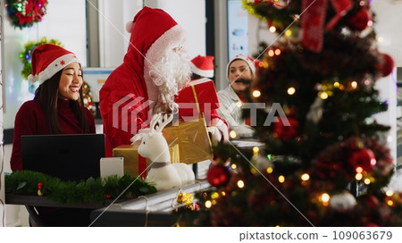 Multiethnic workers receiving gifts from colleague pretending to be Santa in xmas decorated office. Employee wearing costume surprising company personnel with presents during winter holiday season 109063679
