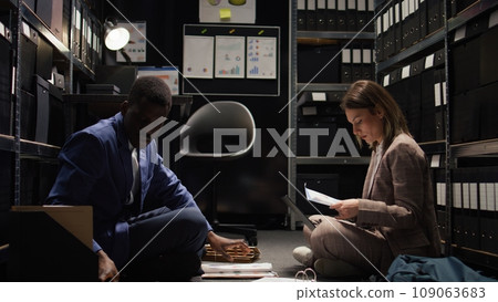 In well-equipped room, multiethnic team of private detectives conduct thorough investigations. Two investigators gather case information, examining witness testimonies, and doing research with laptop. In well-equipped room, multiethnic team of private detectives conduct thorough investigations. Two investigators gather case information, examining witness testimonies, and doing research with laptop. 109063683