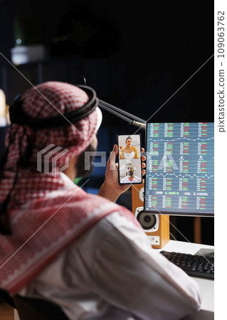 Middle Eastern man engrossed in a video conference call with a coworkers on his cell phone. Muslim guy uses wireless technology for work and research, showcasing his digital communication skills. Middle Eastern man engrossed in a video conference call with a coworkers on his cell phone. Muslim guy uses wireless technology for work and research, showcasing his digital communication skills. 109063762
