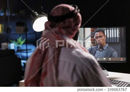 Male African-American and Middle Eastern entrepreneurs engaged in online video conferencing. Muslim man dressed in traditional clothing attentively listening to his coworker on his desktop pc. Male African-American and Middle Eastern entrepreneurs engaged in online video conferencing. Muslim man dressed in traditional clothing attentively listening to his coworker on his desktop pc. 109063767