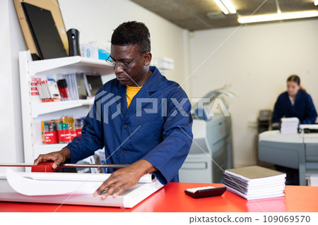 Focused African American man in blue robe uniform trimming the printed work on the cutter in the typography 109069570