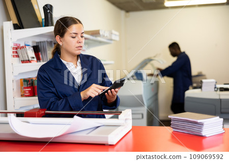 Attentive young woman in a blue uniform calculating the cost of the work done in the printing house Attentive young woman in a blue uniform calculating the cost of the work done in the printing house 109069592