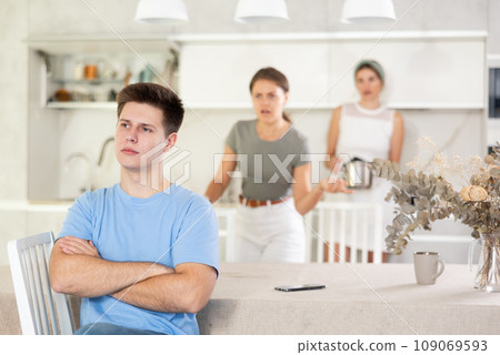 Upset guy sitting in kitchen on background of dissatisfied wife and mother-in-law Upset guy sitting in kitchen on background of dissatisfied wife and mother-in-law 109069593