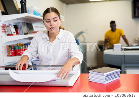 Concentrated young woman in white shirt using paper cutter on the table with planners and calculator in the printer house Concentrated young woman in white shirt using paper cutter on the table with planners and calculator in the printer house 109069612