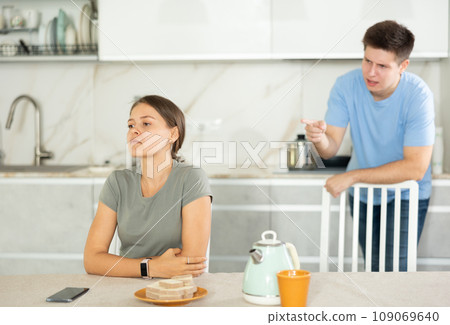 Young girl sitting at the table while her boyfriend is quarreling with her standing behind 109069640
