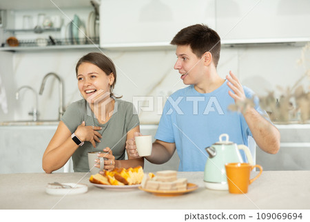 Cheerful young couple drinking tea and talking at kitchen table Cheerful young couple drinking tea and talking at kitchen table 109069694