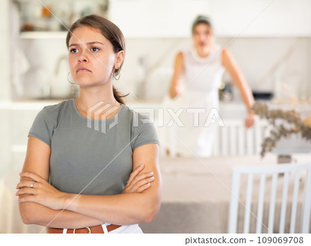 Offended girl sits at table in kitchen and listens to woman scolding her Offended girl sits at table in kitchen and listens to woman scolding her 109069708