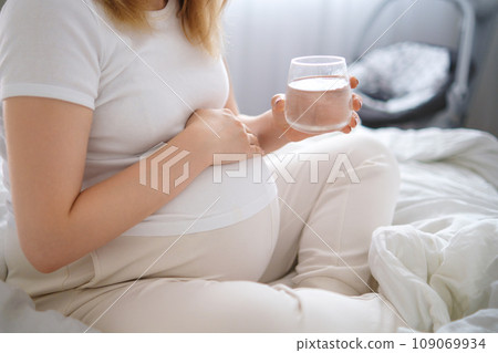 Pregnant woman sits on the bed, tenderly embracing her growing belly and holding glass of water. 109069934
