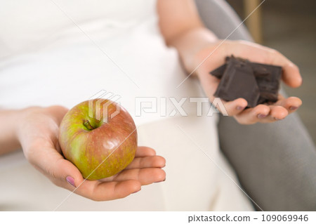 Close up apple and chocolate in pregnant woman hands. Sweets against fruits. Close up apple and chocolate in pregnant woman hands. Sweets against fruits. 109069946