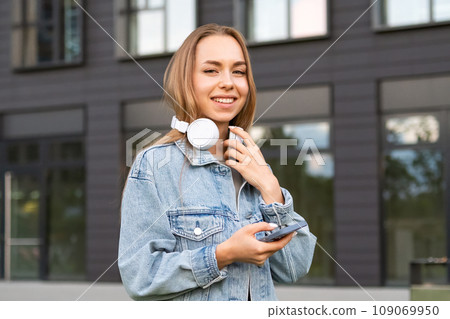 A young woman with a content expression balances headphones on her neck while holding a phone A young woman with a content expression balances headphones on her neck while holding a phone 109069950