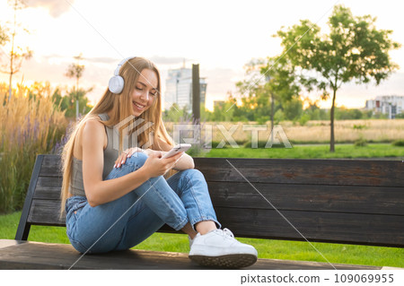 Blonde girl sits cross legged on a bench and listens to music via headphones in the park at sunset. Blonde girl sits cross legged on a bench and listens to music via headphones in the park at sunset. 109069955