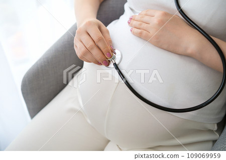 Pregnant woman, dressed in a light outfit listening to her stomach with a stethoscope.  109069959
