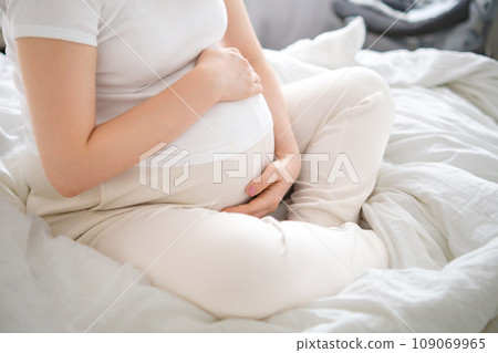 Pregnant woman takes a seat on the bed, tenderly embracing her growing belly 109069965