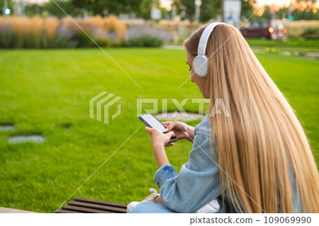 Blonde girl sits with her back in headphones and uses a mobile phone in a clearing in the park. Blonde girl sits with her back in headphones and uses a mobile phone in a clearing in the park. 109069990