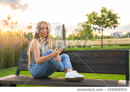 Freelancer enjoys music via headphones sitting on the bench in the park. Freelancer enjoys music via headphones sitting on the bench in the park. 109069993