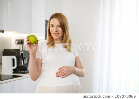 Beautiful blonde pregnant woman holding a pear and tough her belly standing in the kitchen. Beautiful blonde pregnant woman holding a pear and tough her belly standing in the kitchen. 109070069