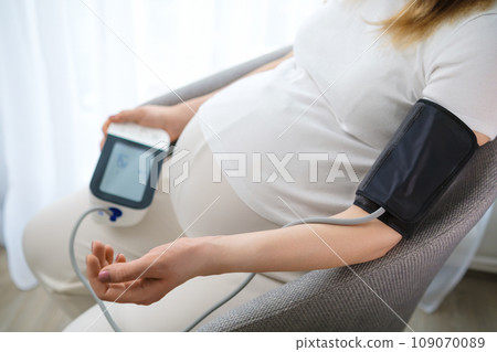 Future mother controls blood pressure with measuring instrument. Woman sits comfortably on chair looking at device expecting results crop body 109070089