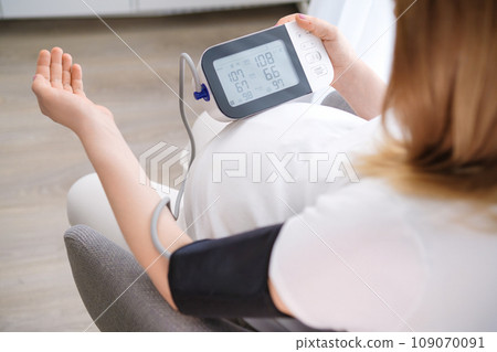 Pregnant woman sits on gray chair controlling blood pressure with measuring instrument. Lady hand rests on table ensuring accurate readings crop body 109070091