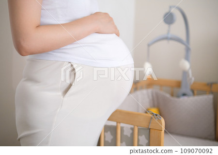 Pregnant woman stands near baby bed with hanging toys. Woman fingers trace contours of crib simultaneously caressing belly in anticipation of baby Pregnant woman stands near baby bed with hanging toys. Woman fingers trace contours of crib simultaneously caressing belly in anticipation of baby 109070092