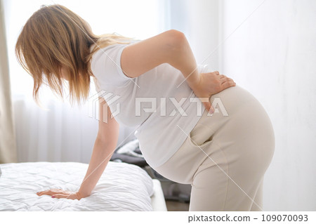 Expectant mother leans on soft bed in bedroom holding hand on hip. Future mother slowly rocks body alleviating back pain during pregnancy crop body Expectant mother leans on soft bed in bedroom holding hand on hip. Future mother slowly rocks body alleviating back pain during pregnancy crop body 109070093