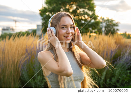 Blonde young woman listening music using headphones while resting in the park. Blonde young woman listening music using headphones while resting in the park. 109070095
