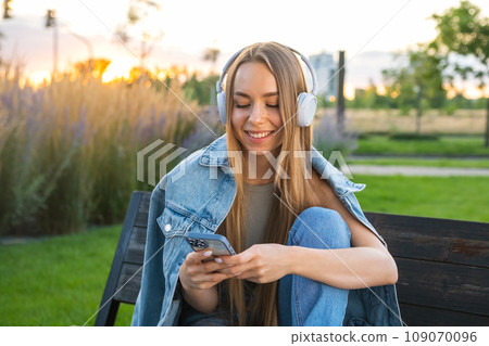 Young blonde woman in denim uses a mobile phone and listen to music sitting on the bench in the park Young blonde woman in denim uses a mobile phone and listen to music sitting on the bench in the park 109070096