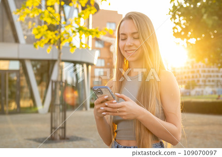 Happy young woman with long blonde hair holds a mobile phone standing in the street. Happy young woman with long blonde hair holds a mobile phone standing in the street. 109070097
