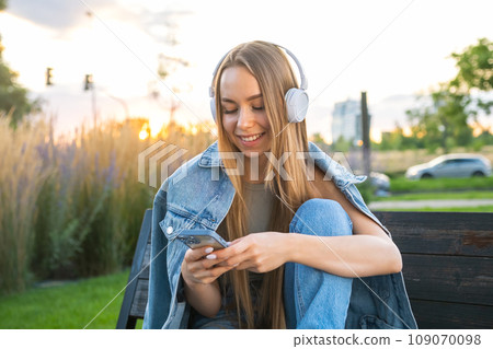 Female student with long blonde hair uses smart phone and listen to music after study sitting on the bench at sunset. Female student with long blonde hair uses smart phone and listen to music after study sitting on the bench at sunset. 109070098
