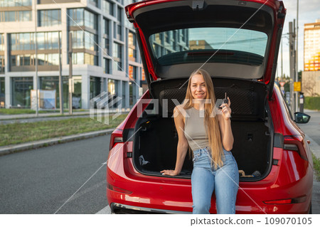 Young blonde woman sitting in the trunk of new red car and holding keys. Young blonde woman sitting in the trunk of new red car and holding keys. 109070105