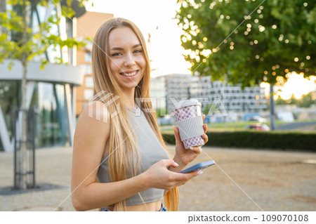 Cheerful young woman smiles at camera holding a cup and smart phone in her hands, copy space 109070108