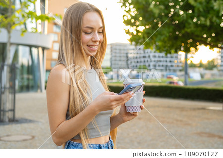 Blonde hair woman smiles at phone screen texting message standing in the street at sunset.  109070127