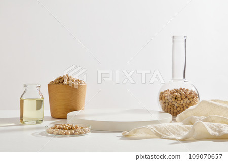 A petri dish, flat-bottom florence flask and wooden bowl of soybeans arranged with a jar of soybean oil and a podium. White background. Minimal scene with geometric podium platform 109070657