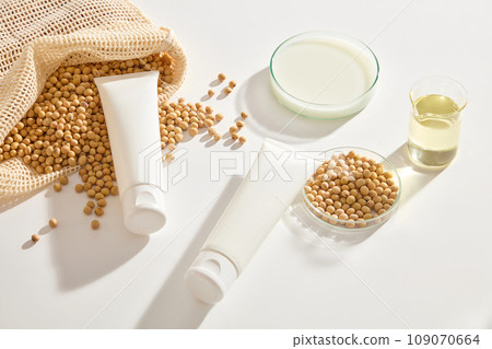 Blank label tubes in white color displayed with a mesh bag of soybeans and glassware filled with milk and oil. Soybean is associated with reducing the visible effects of skin aging 109070664