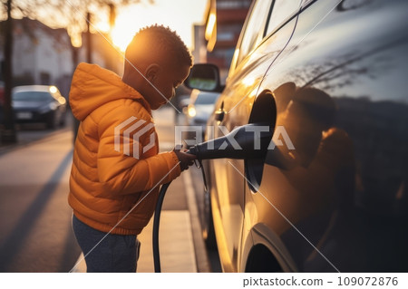 Little African American boy attaching power cable to electric vehicle at charging station. Driver's young helper inserts electrical connector to charge battery. Electric vehicle charging plug. Little African American boy attaching power cable to electric vehicle at charging station. Driver's young helper inserts electrical connector to charge battery. Electric vehicle charging plug. 109072876
