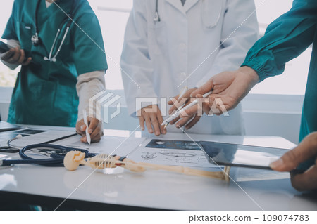 Multiracial medical team having a meeting with doctors in white lab coats and surgical scrubs seated at a table discussing a patients records Multiracial medical team having a meeting with doctors in white lab coats and surgical scrubs seated at a table discussing a patients records 109074783