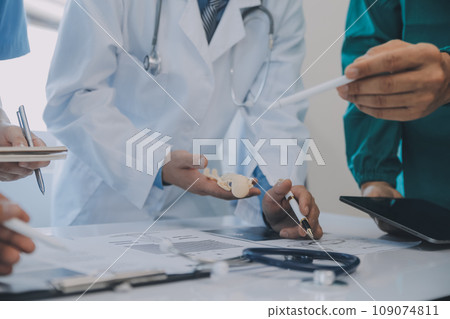 Multiracial medical team having a meeting with doctors in white lab coats and surgical scrubs seated at a table discussing a patients records 109074811