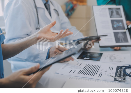Multiracial medical team having a meeting with doctors in white lab coats and surgical scrubs seated at a table discussing a patients records 109074828