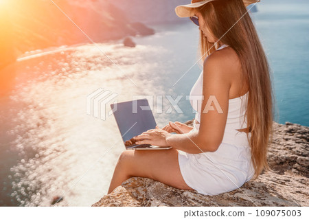 Digital nomad, woman in the hat, a business woman with a laptop sits on the rocks by the sea during sunset, makes a business transaction online from a distance. Freelance, remote work on vacation. 109075003