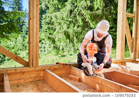 Father with toddler son constructing wooden frame house near forest. Male worker teaching his son how to cut board using electric saw on construction site, wearing helmets and overalls on sunny day. 109079283