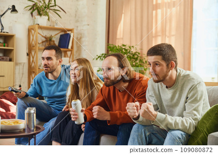Attractive young people, man and woman sitting in front of TV and attentively watching show, football match and eating snacks with alcohol drinks. 109080872
