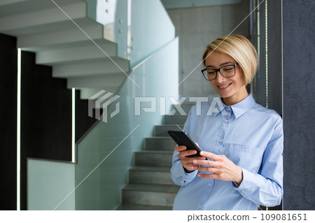 Young happy woman inside office at workplace, blonde businesswoman using phone, smiling reading news on smartphone, standing near window, typing text message call. Young happy woman inside office at workplace, blonde businesswoman using phone, smiling reading news on smartphone, standing near window, typing text message call. 109081651