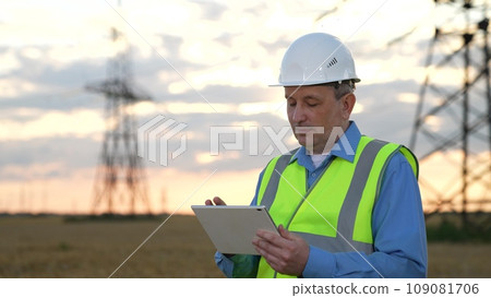 Mature engineer inputs data on tablet and looks back at power transmission line Mature engineer inputs data on tablet and looks back at power transmission line 109081706