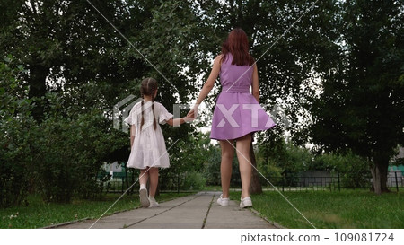 Young mother holds little daughter hand walking on path across green summer park Young mother holds little daughter hand walking on path across green summer park 109081724