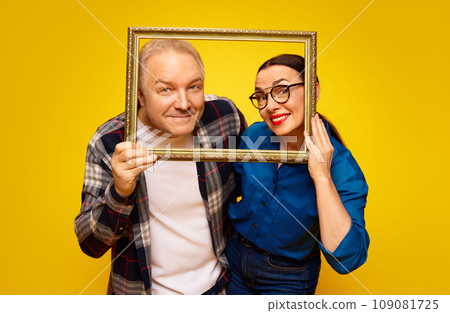 Portrait of happy, loving, senior couple, man and woman posing with picture frame against yellow studio background. Valentine's Day Portrait of happy, loving, senior couple, man and woman posing with picture frame against yellow studio background. Valentine's Day 109081725