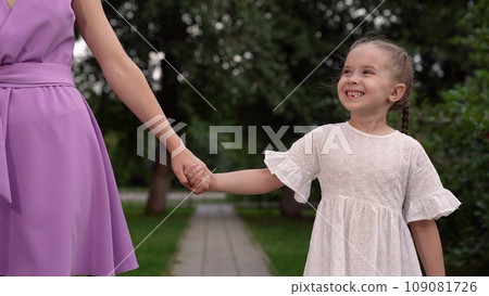 Joyful little daughter smiles and looks at mother holding hands at walk in park Joyful little daughter smiles and looks at mother holding hands at walk in park 109081726