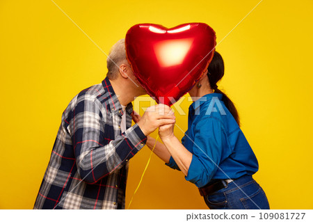 Senior couple, wife and husband standing behind red balloon in heart shape and kissing against yellow studio background. Valentine's Day Senior couple, wife and husband standing behind red balloon in heart shape and kissing against yellow studio background. Valentine's Day 109081727