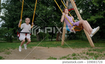 Cute little daughter waves hand to mother playing swings together on playground Cute little daughter waves hand to mother playing swings together on playground 109081728