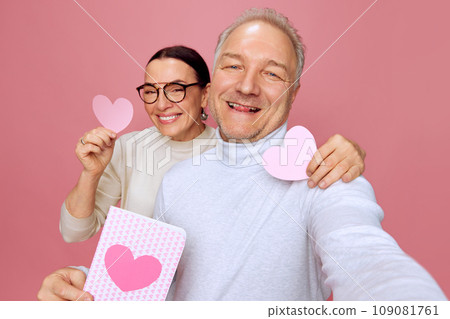 Selfie time. Happy, smiling senior man and woman taking holiday photo with paper heart postcards against pink studio background. Valentine's Day Selfie time. Happy, smiling senior man and woman taking holiday photo with paper heart postcards against pink studio background. Valentine's Day 109081761