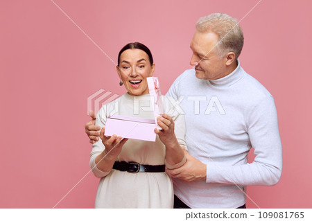 Senior beautiful woman opening present with happiness and excitement against pink studio background. Man surprising wife. Valentine's Day Senior beautiful woman opening present with happiness and excitement against pink studio background. Man surprising wife. Valentine's Day 109081765